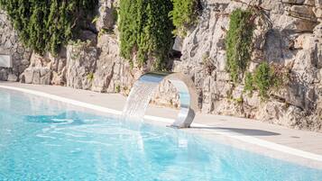 A swimming pool with clear blue water, a stone wall, and greenery.