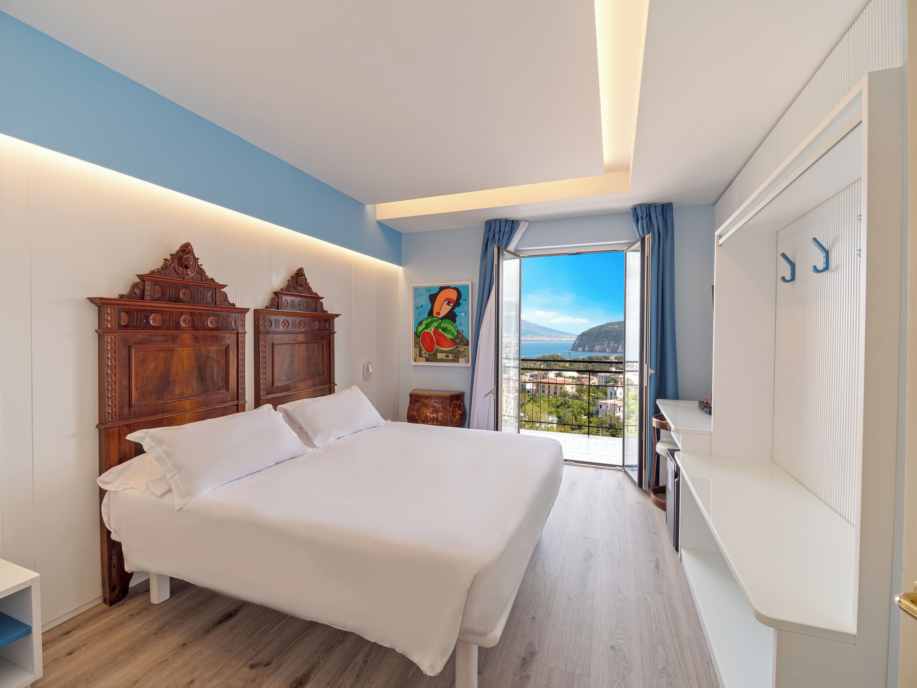 A bedroom with a large bed, wooden headboard, a painting, a balcony with a view, and a white wardrobe.