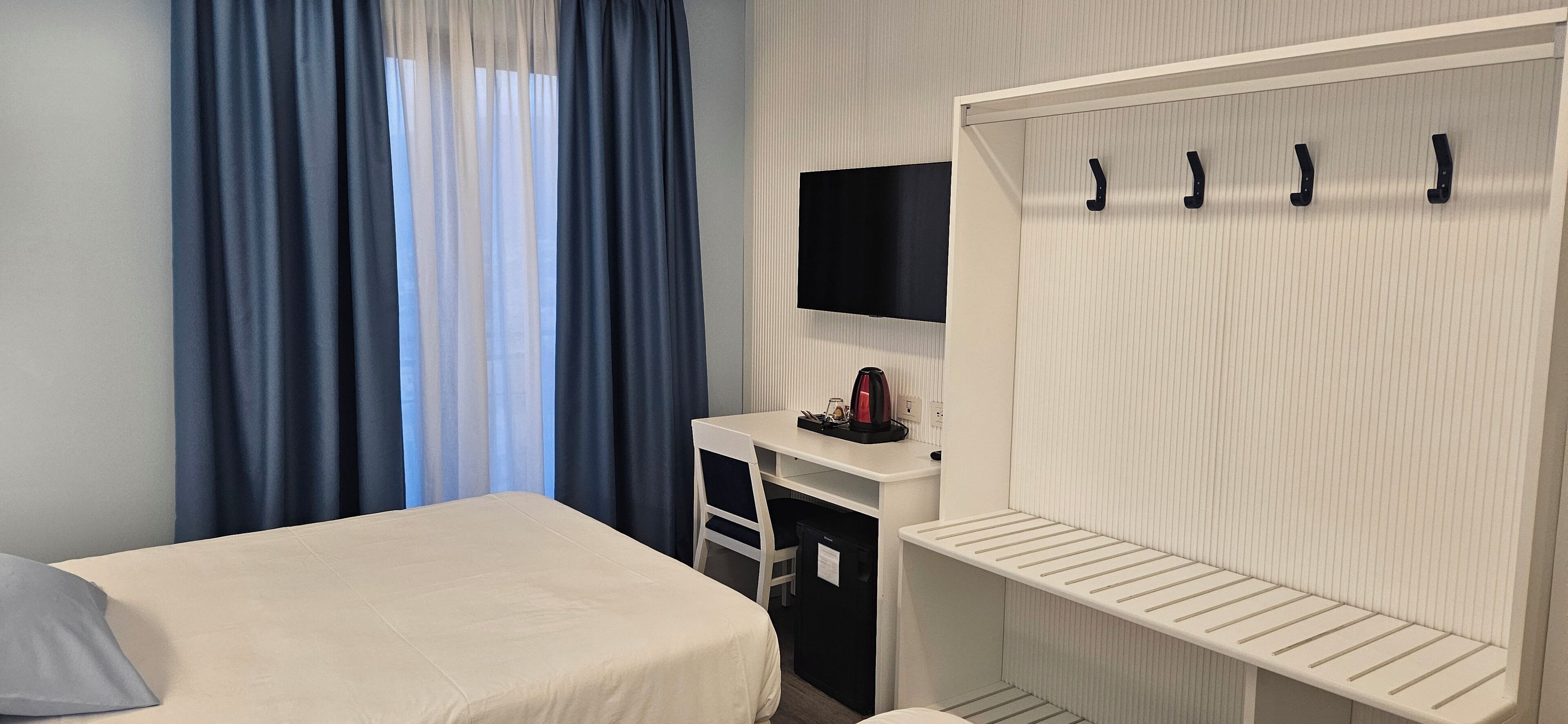 A hotel room with a bed, a desk with a flat-screen TV, a wardrobe with hooks, and a window with blue curtains.