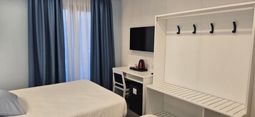 A hotel room with a bed, a desk with a flat-screen TV, a wardrobe with hooks, and a window with blue curtains.