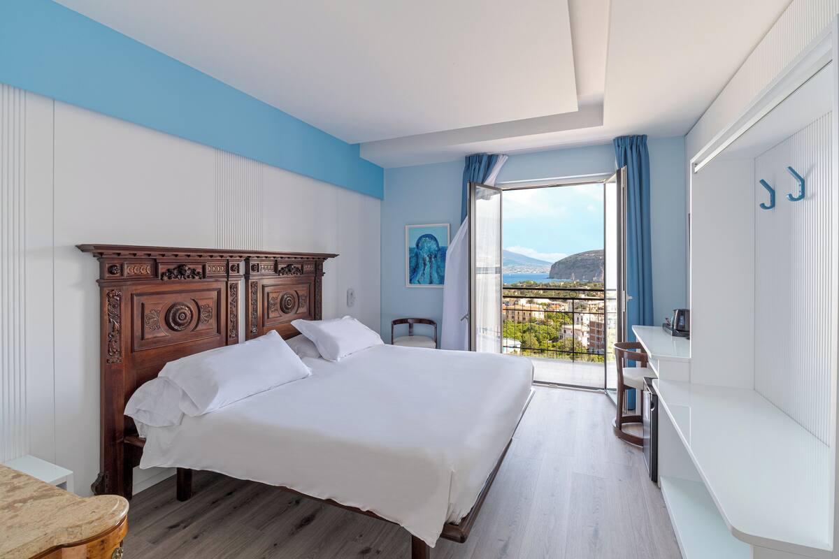 A bedroom with a large bed, a wooden headboard, a view of the city through a balcony, and a bathroom with a white sink and mirror.
