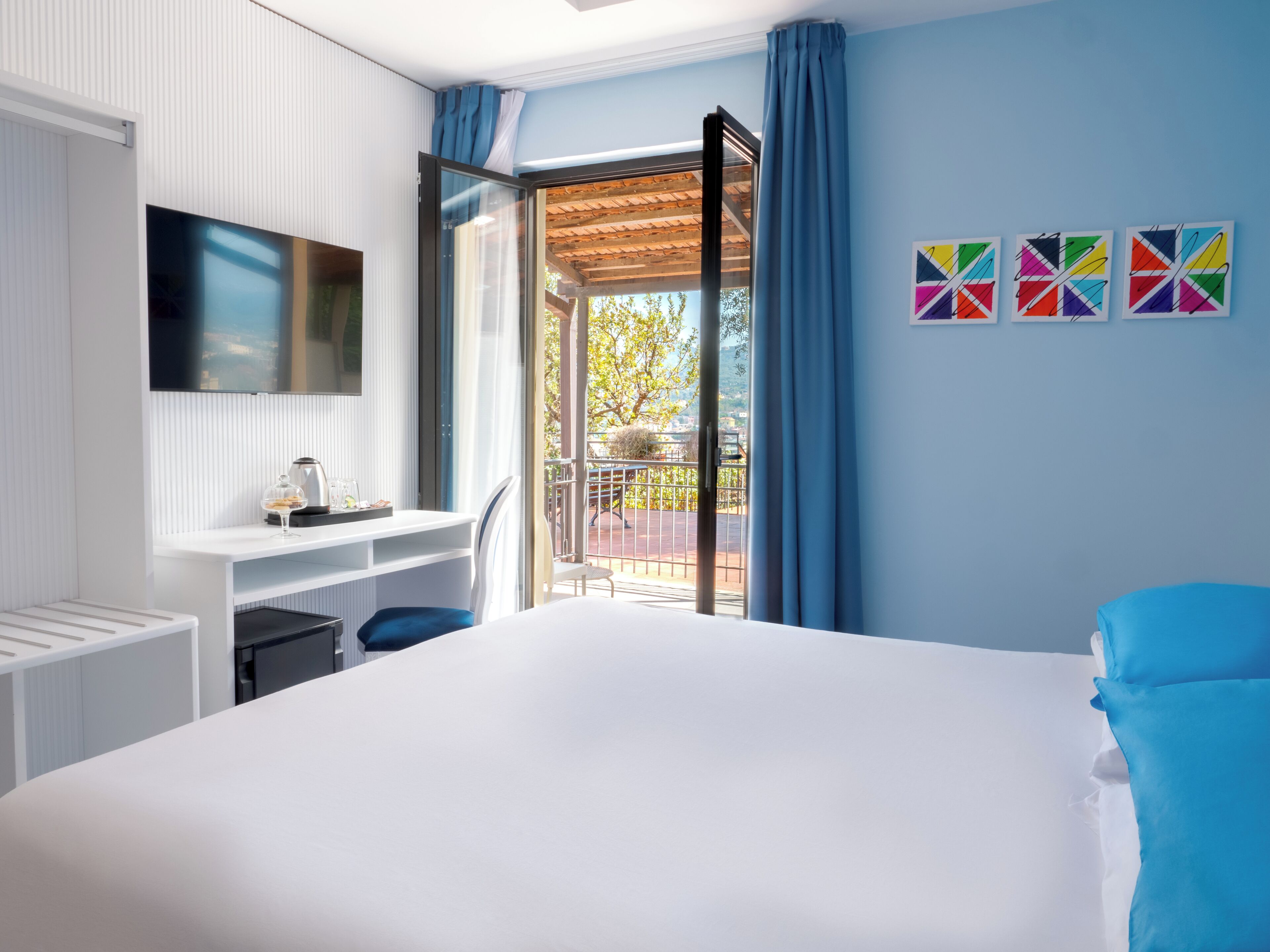 Una habitación de hotel moderna con una cama grande, un escritorio con televisor de pantalla plana, un balcón con cortinas azules y obras de arte coloridas en la pared.