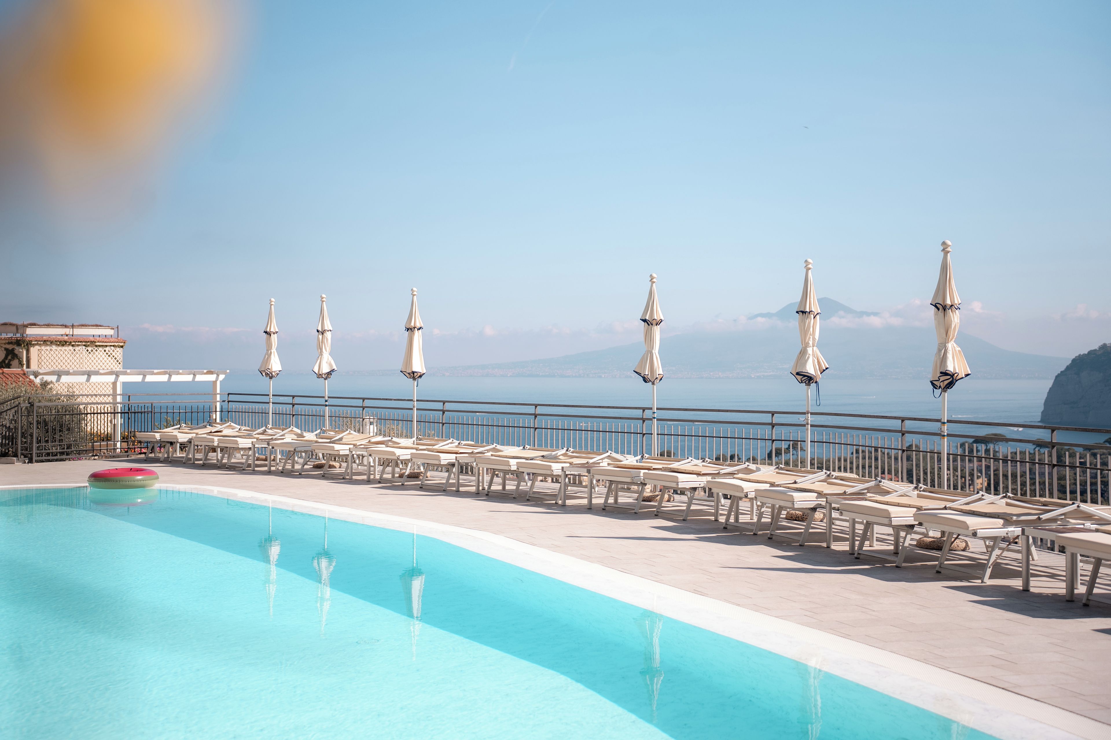 Zona de piscina con sillones de descanso y sombrillas, con vistas a una masa de agua y montañas distantes.