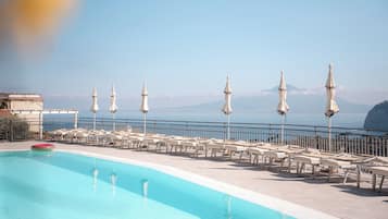 Zona de piscina con sillones de descanso y sombrillas, con vistas a una masa de agua y montañas distantes.