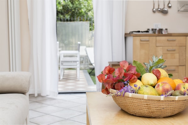 Una cesta de mimbre llena de frutas y flores sobre una mesa en una habitación bien iluminada, con vistas a un patio.