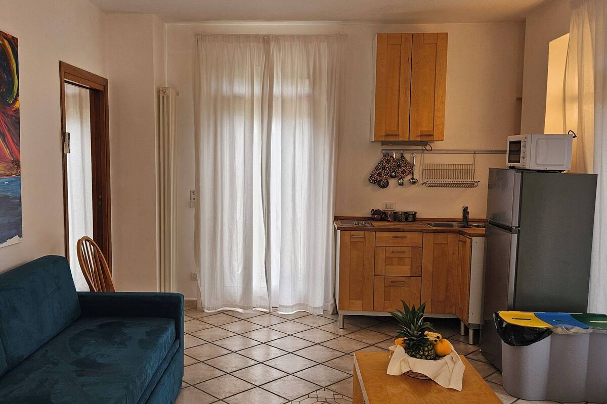 A compact kitchen with wooden cabinets, a microwave, and a refrigerator. There's a dining table with fruits and a chair. A colorful painting adorns the wall.