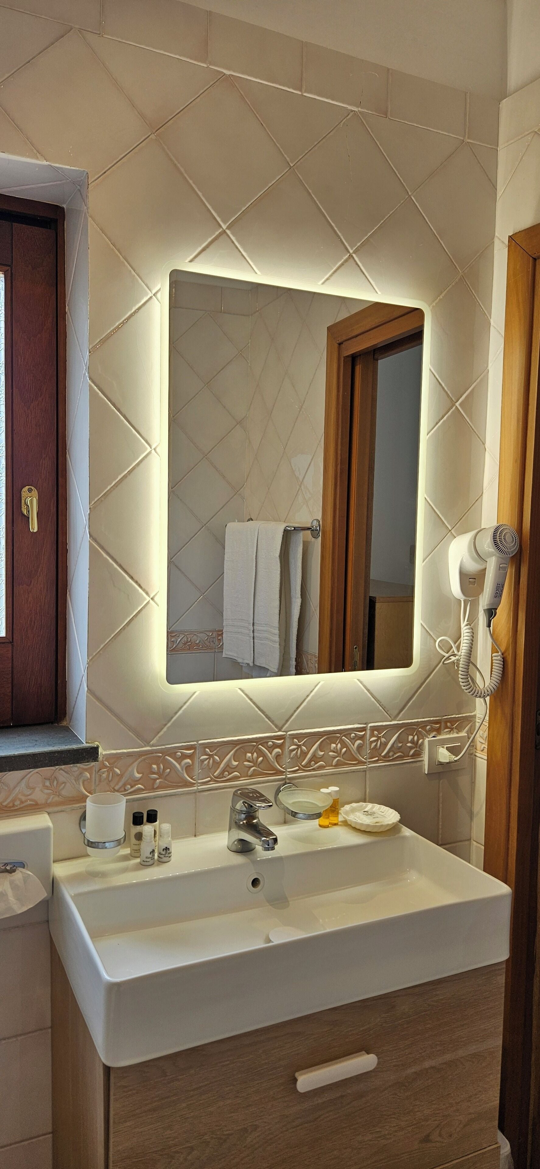 Casa de banho com um espelho grande, secador de cabelo, lavatório com artigos de higiene e porta de madeira.