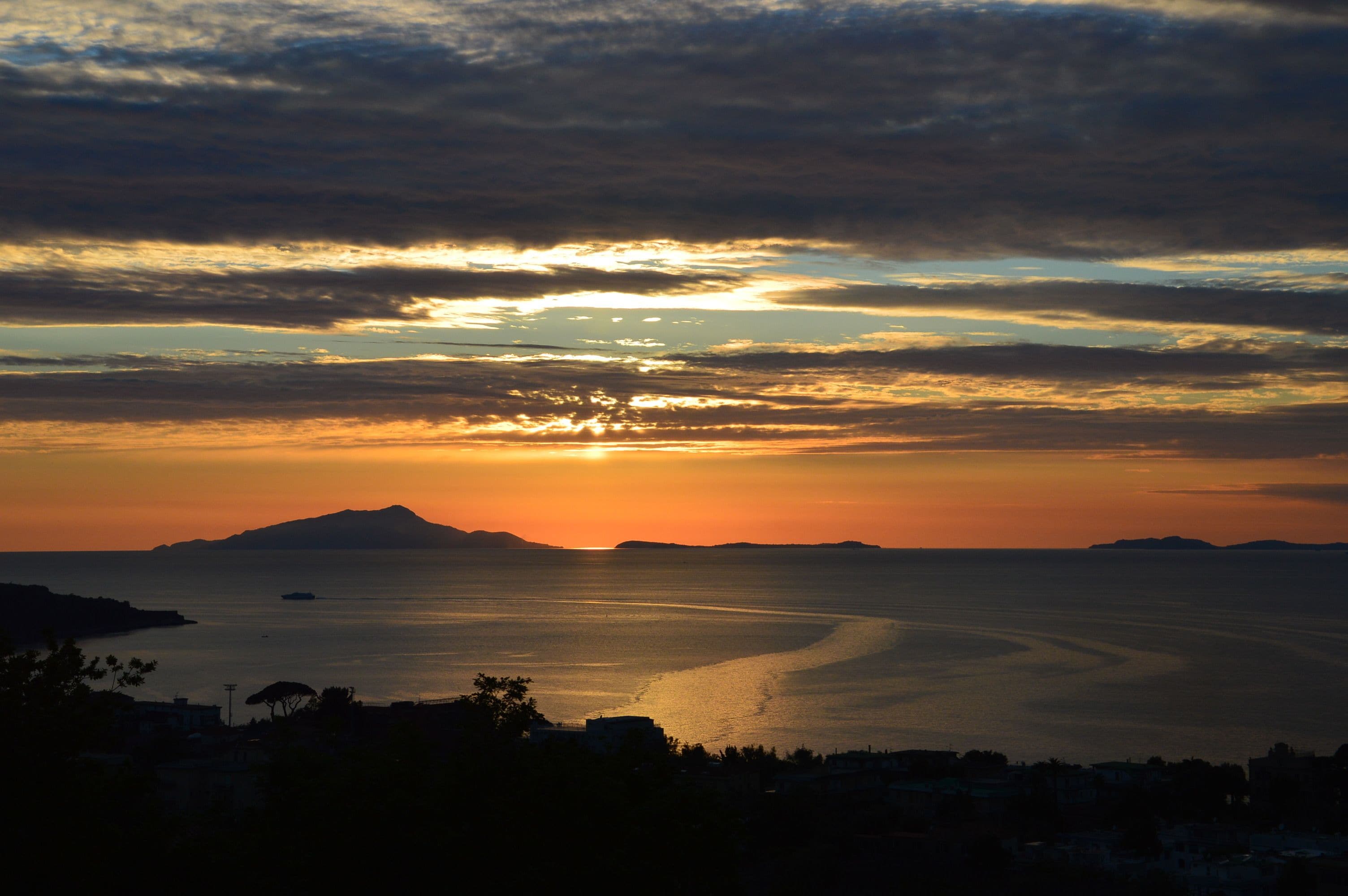 Un coucher de soleil sur la côte, avec une mer calme, des montagnes lointaines et la silhouette d’une ville.