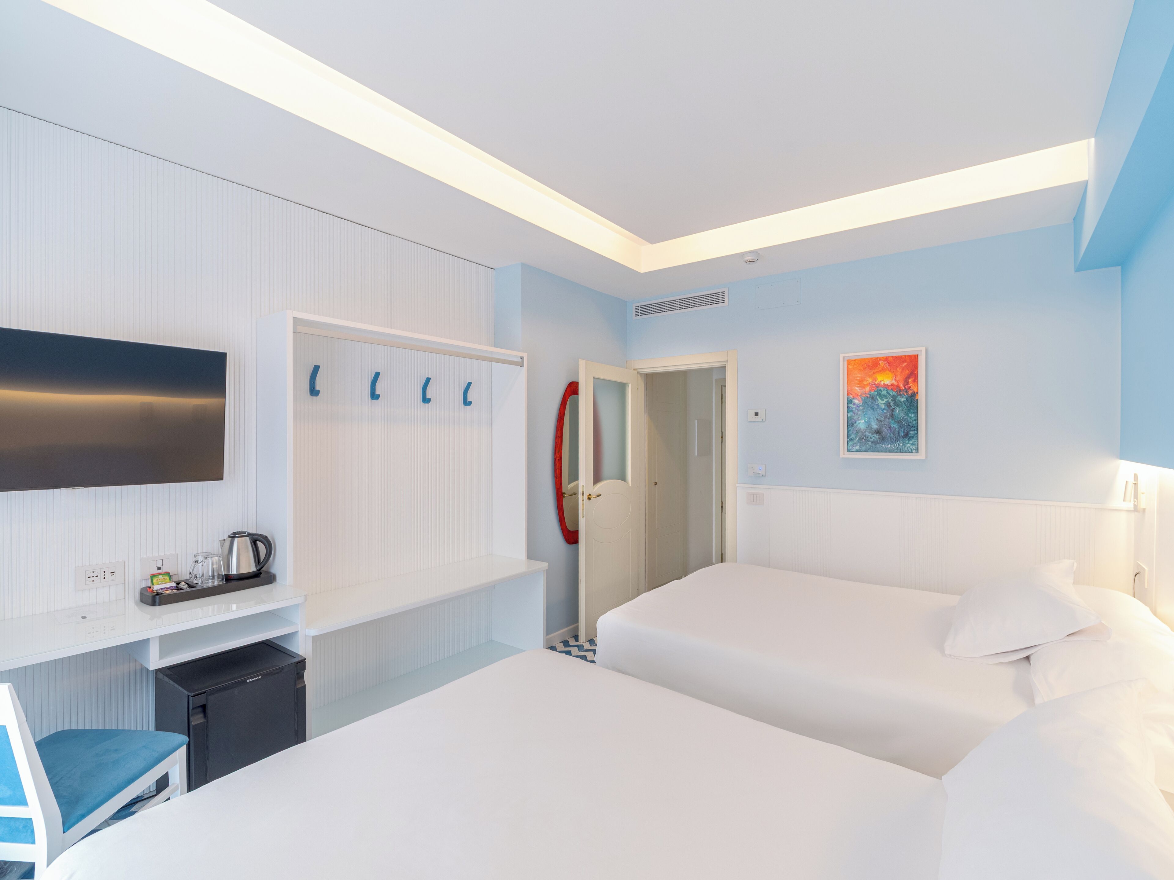 Camera d'albergo con TV a schermo piatto, scrivania con bollitore, mini-frigo, letto con lenzuola bianche e porta che dà accesso al bagno.