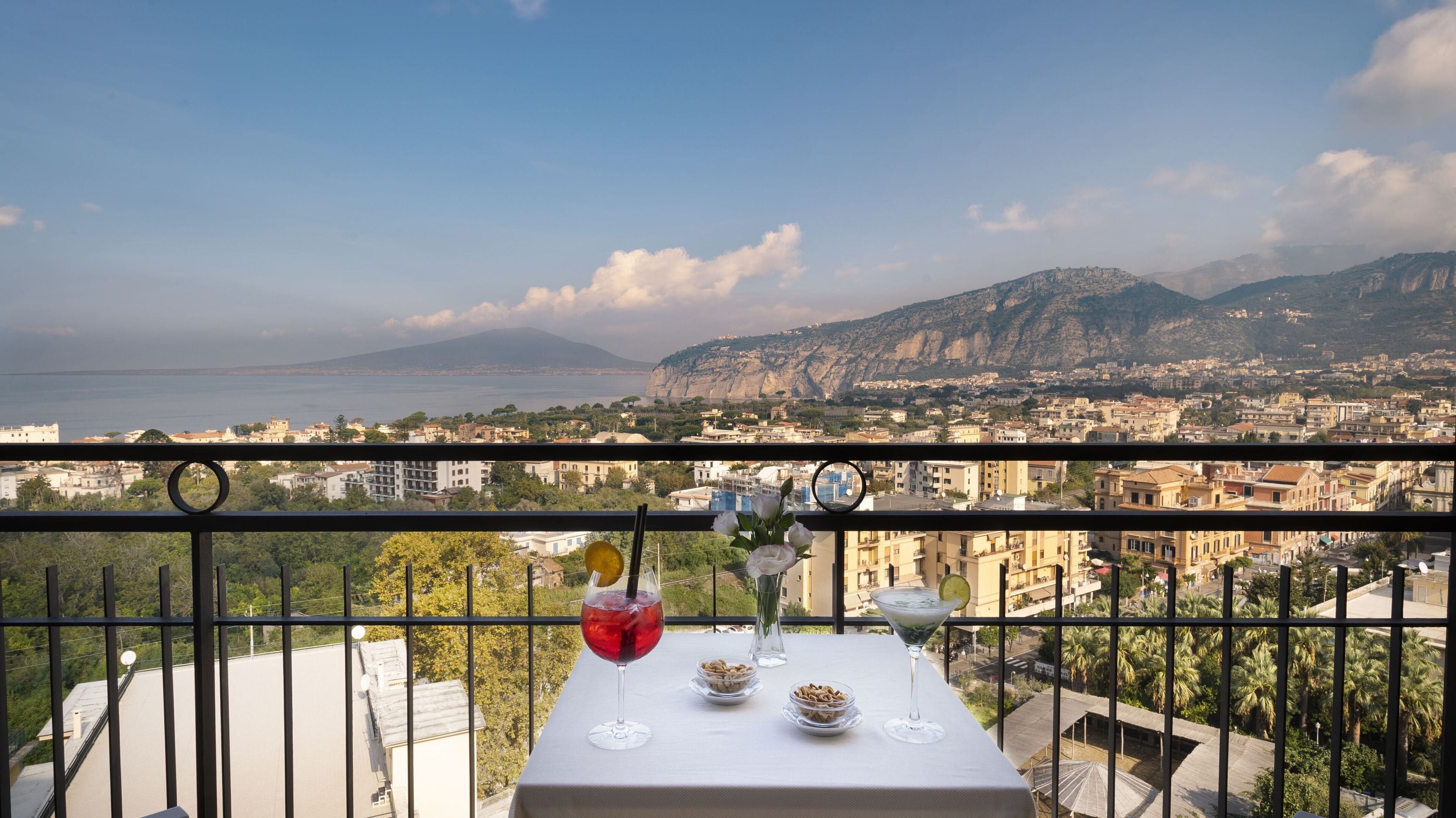 Un balcón con una mesa dispuesta para dos personas, con vistas a una ciudad costera y montañas al fondo.