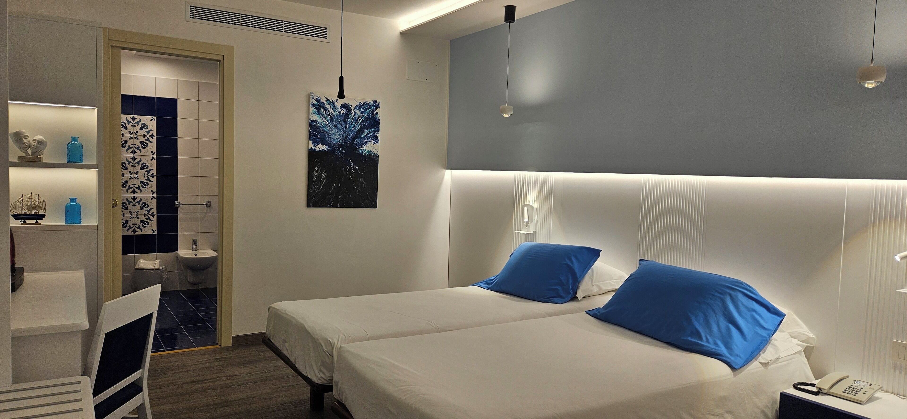 Une chambre d’hôtel avec deux lits, une table de chevet avec un téléphone, une lampe fixée au mur et une salle de bain avec un lavabo et des toilettes.