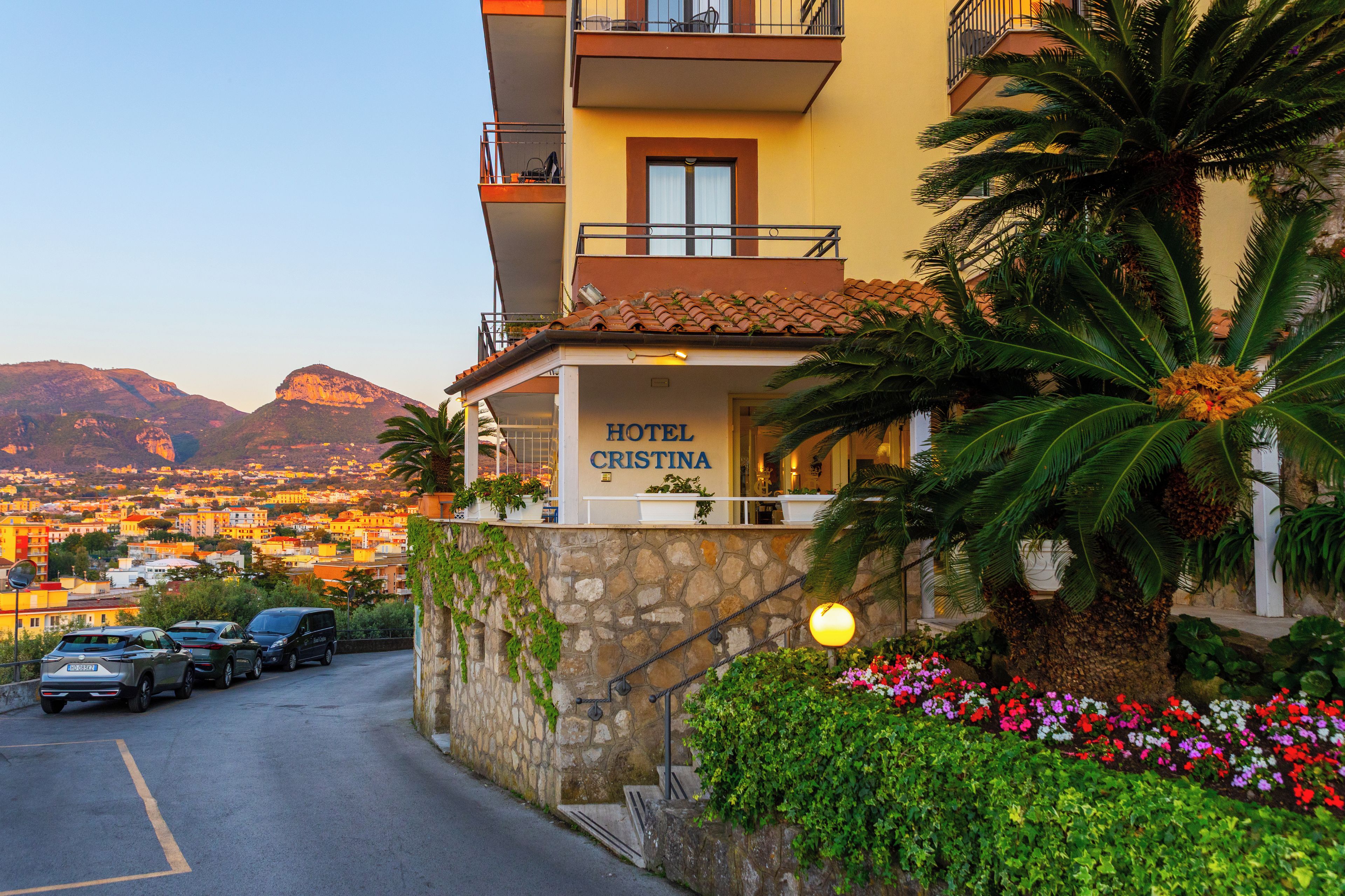 A hotel named 'HOTEL CRISTINA' with a stone facade, a balcony, and a view of a mountainous landscape.