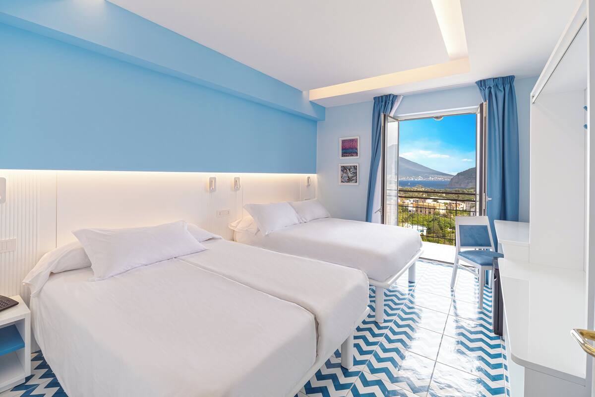 A hotel room with two beds, a view of the city, and a balcony with a chair.