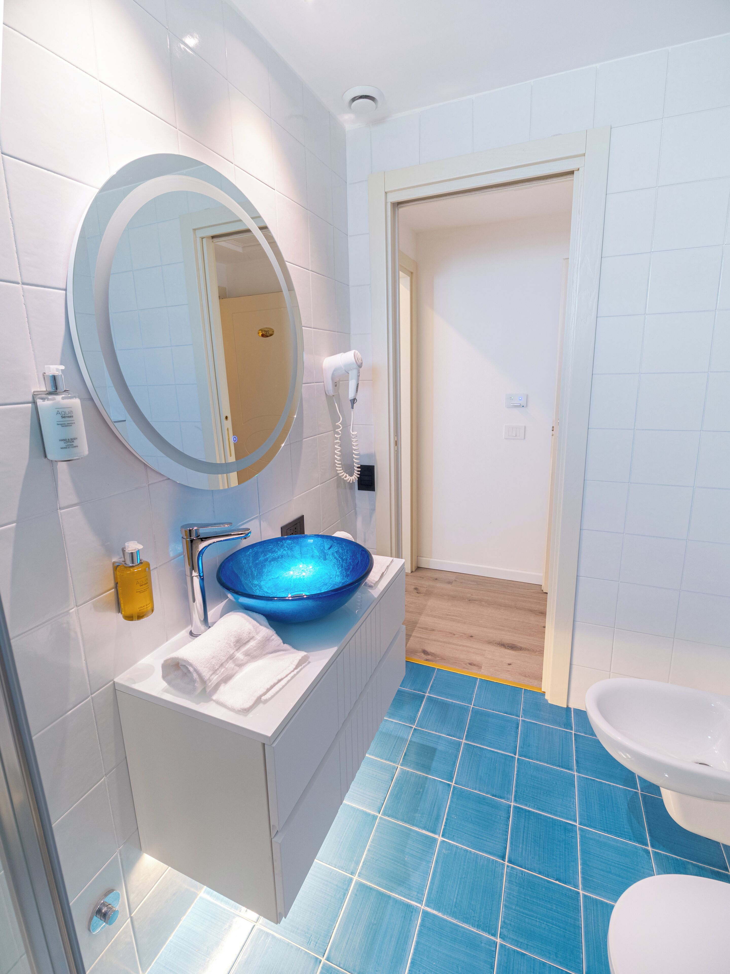 Un baño con piso de azulejos azules, un lavamanos blanco con lavabo azul, un espejo redondo y un secador de pelo en la pared.