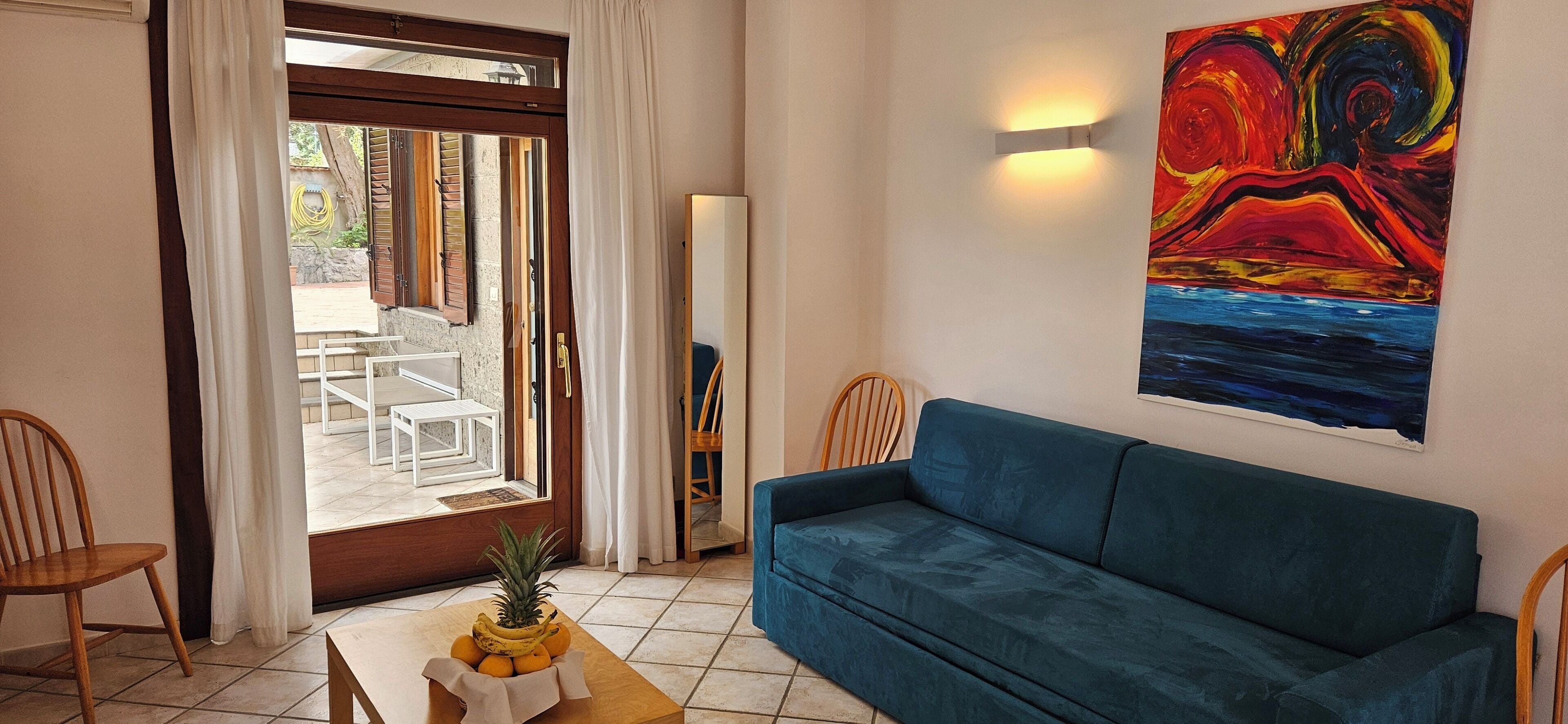 A living room with a blue sofa, a table with fruit, a painting, and a view through a glass door.