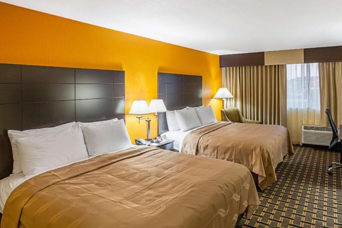 A hotel room with two beds, a nightstand, a lamp, an air conditioning unit, and a window with curtains.