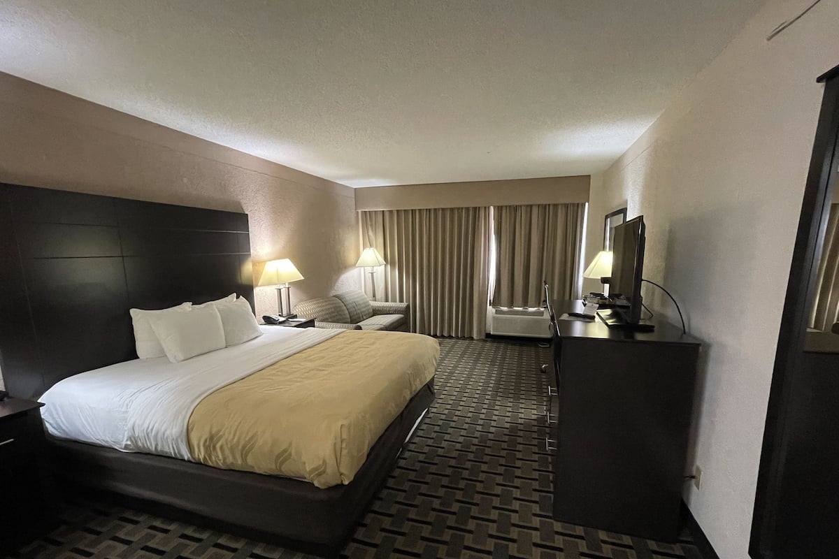 Quarto de hotel com uma cama grande, duas mesas de cabeceira com luminárias, uma escrivaninha com computador e uma televisão.