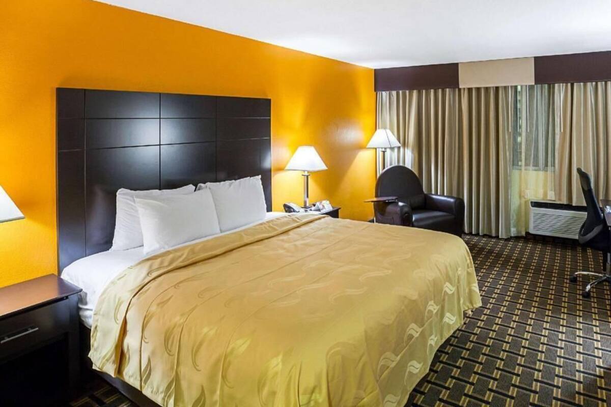 Quarto de hotel com uma cama grande, duas luminárias de cabeceira, uma cadeira, uma escrivaninha e uma janela com cortinas.