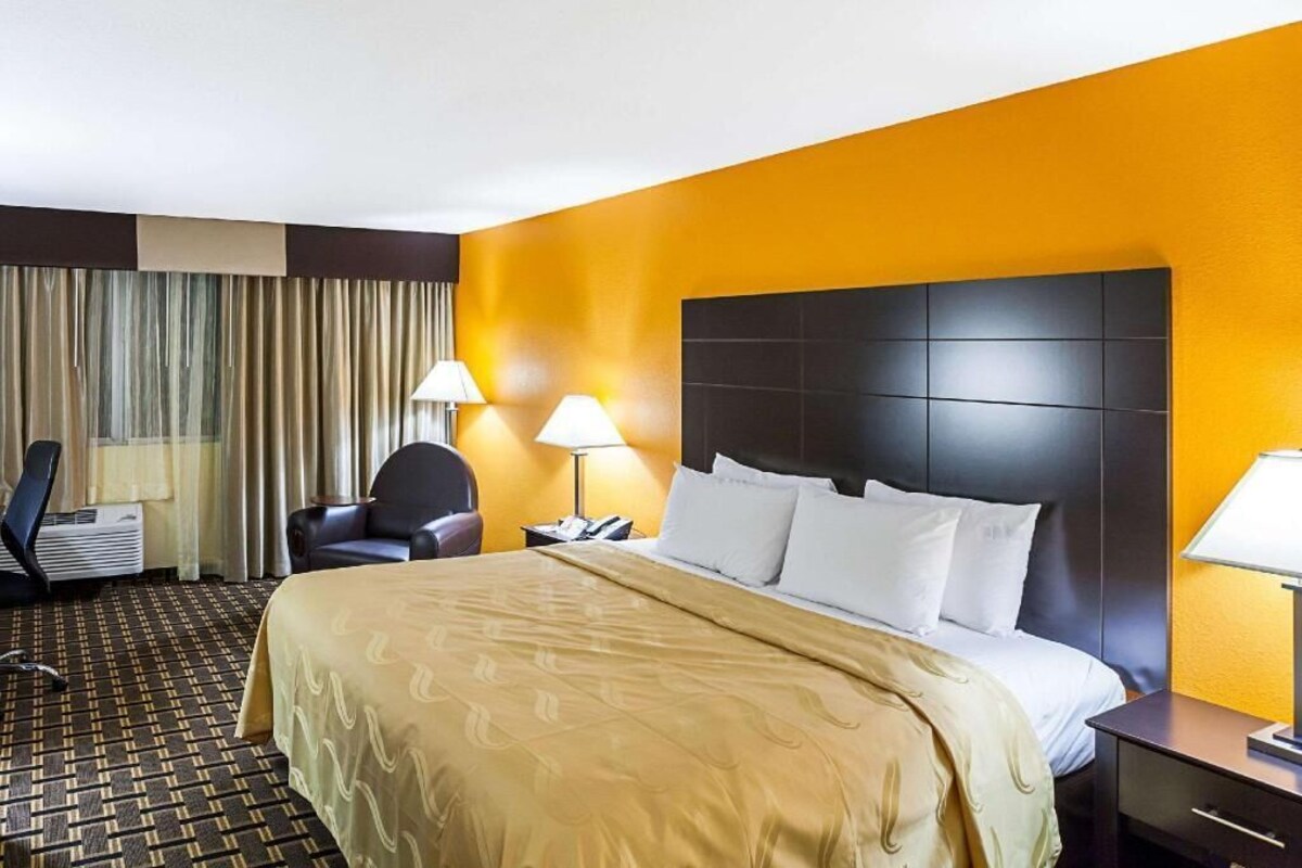 A hotel room with a bed, two bedside lamps, a chair, a desk, and a wall-mounted air conditioner.