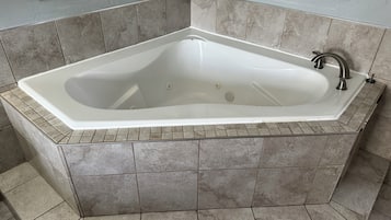 Una bañera de esquina con revestimiento blanco, paredes con azulejos y un espejo en esquina.