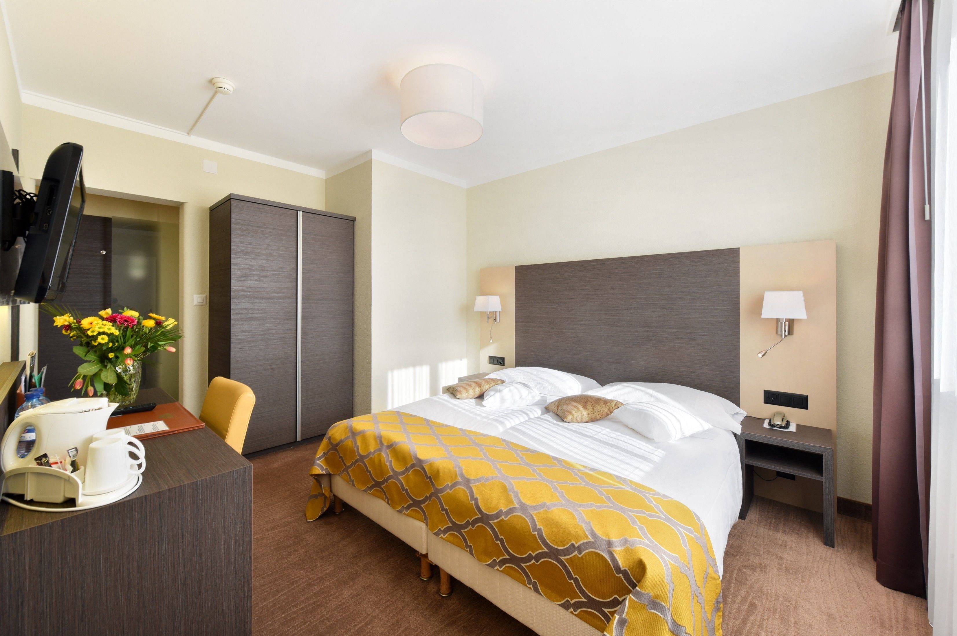 A hotel room with a bed, bedside tables, a desk with a chair, a TV, and a vase with flowers.