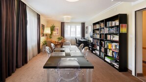 A modern hotel room with a large dining table, clear chairs, a bookshelf, and a desk with a chair.