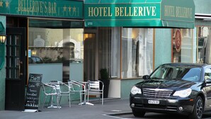 A green awning with 'Hotel Bellerive' written on it, a black car parked in front, and a signboard with 'Bar 99' and 'Notre Dame' visible.