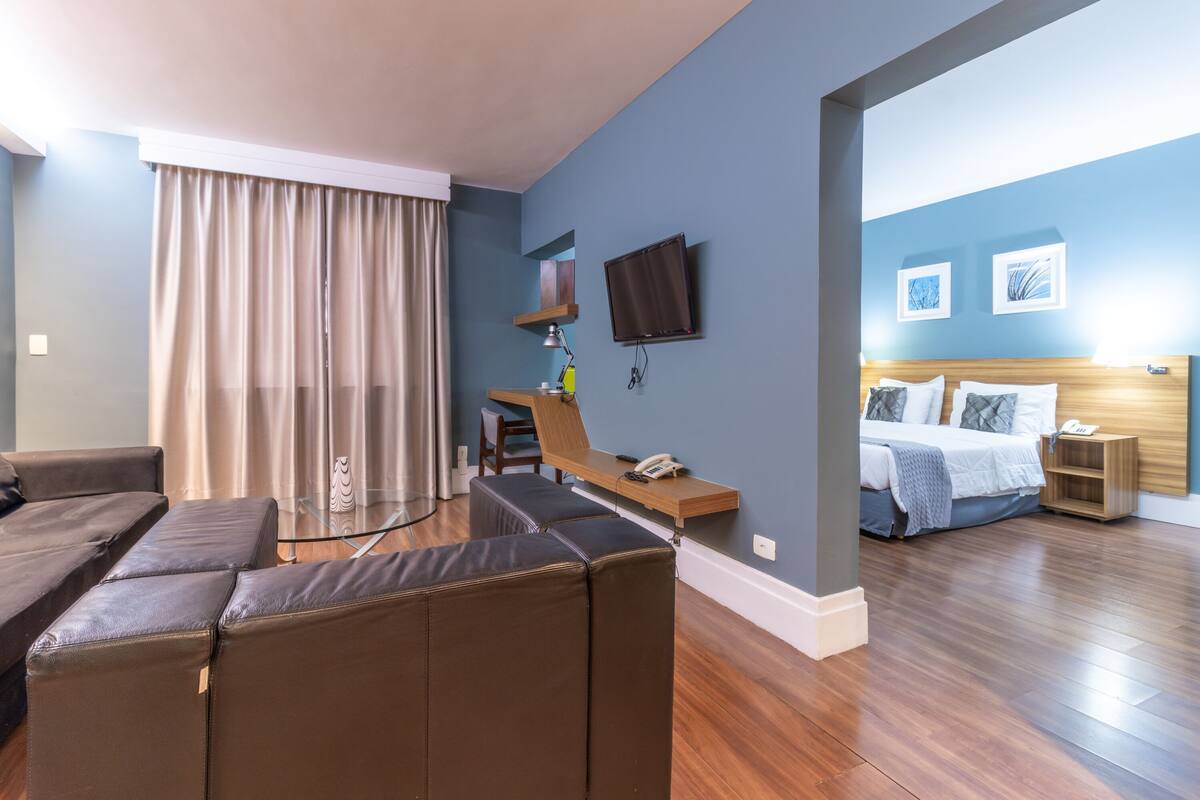 Quarto de hotel moderno com sofá, escrivaninha com luminária, TV de tela plana, cama com travesseiros e obras de arte na parede.