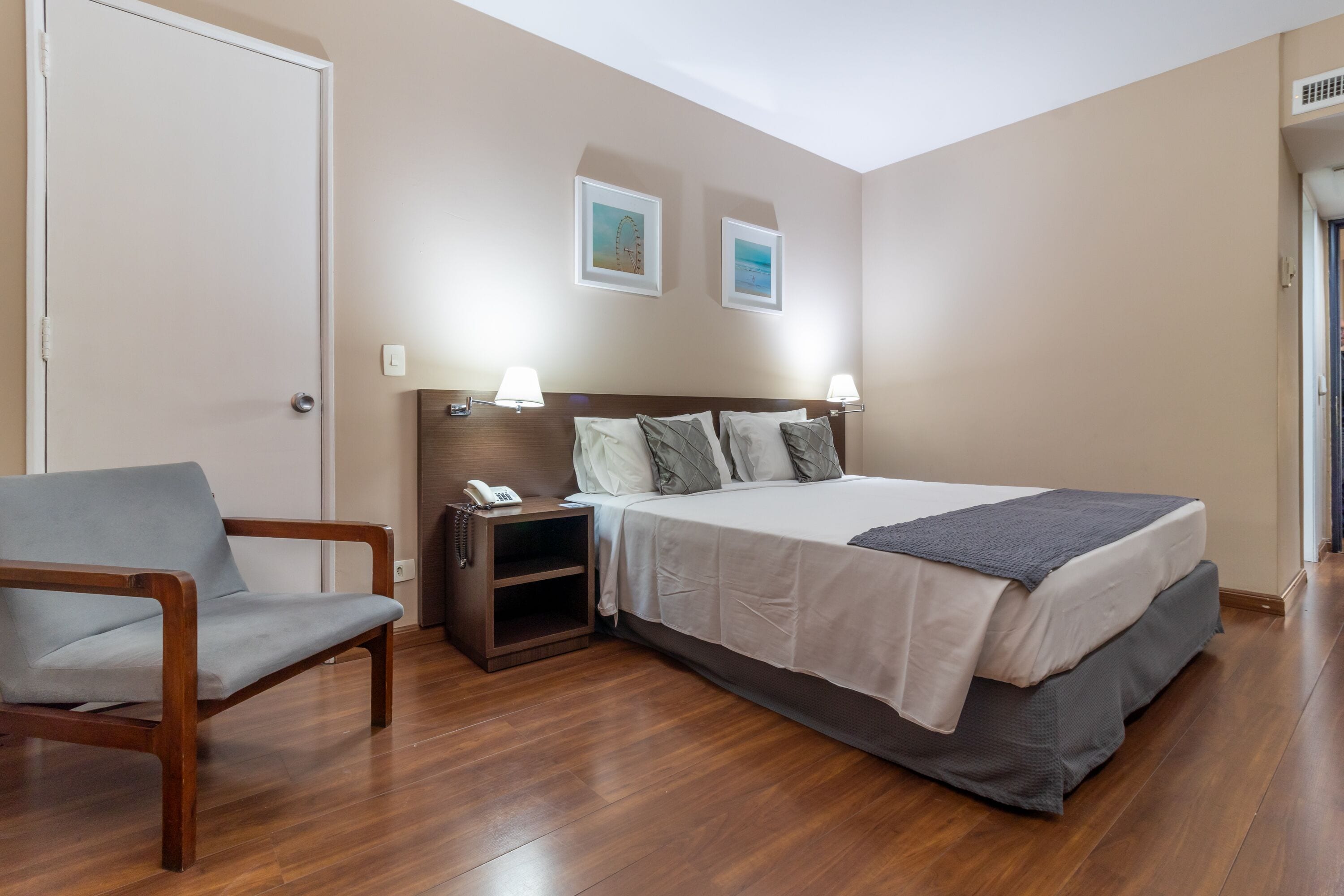A hotel room with a bed, a chair, a nightstand with a phone, and two framed pictures on the wall.