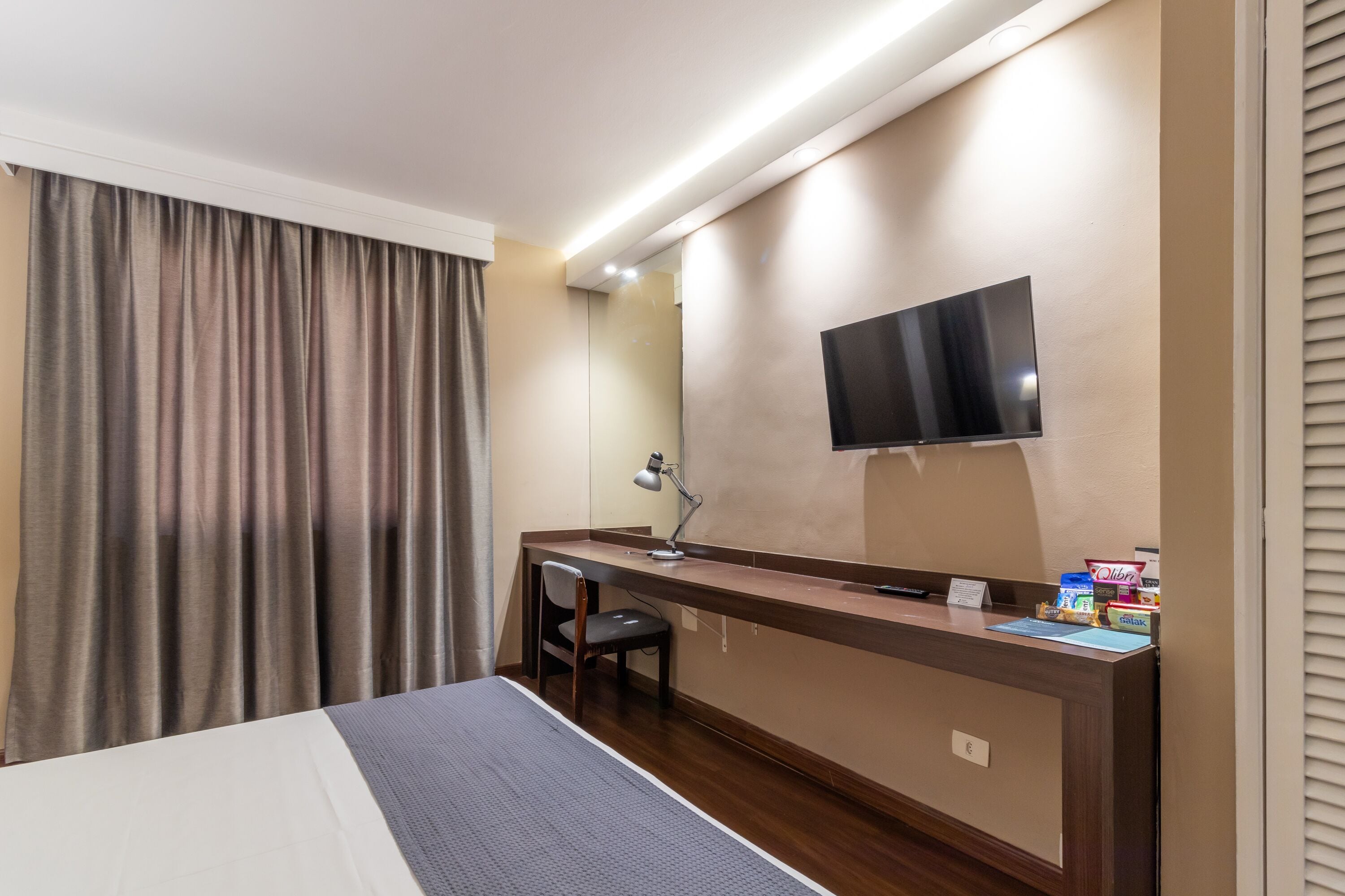 A hotel room with a bed, a desk, a chair, a TV mounted on the wall, and a curtain.