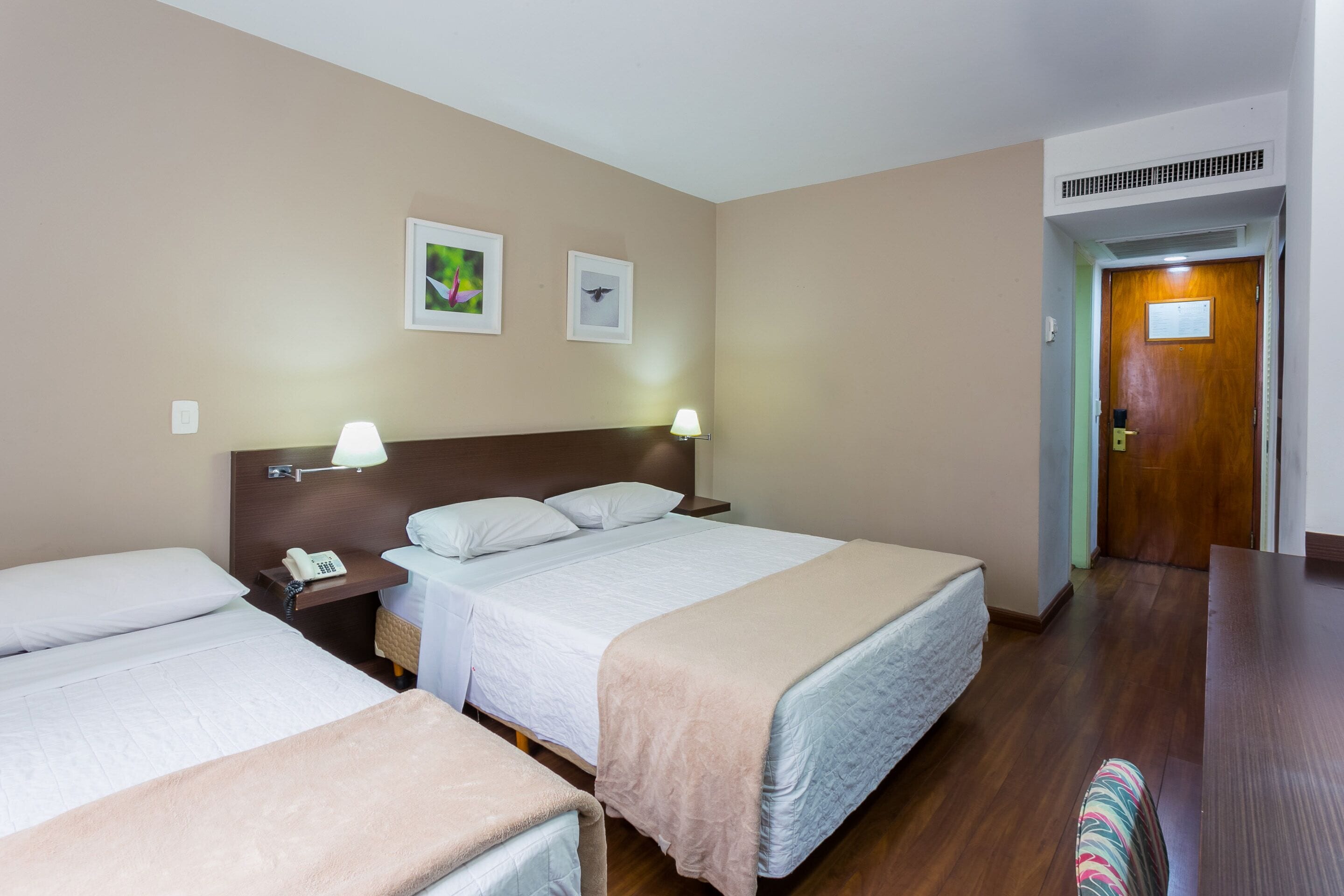 Une chambre d’hôtel avec deux lits, une tête de lit en bois, des lampes de chevet, un téléphone et des tableaux encadrés au mur.