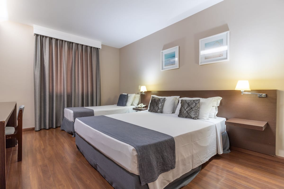 Quarto de hotel com duas camas, uma mesa, uma cadeira, um abajur e um quadro na parede.