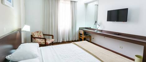Habitación de hotel con cama, una silla, escritorio, televisor y ventana con cortinas.