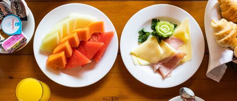 A breakfast setting with plates of fruit, cheese, ham, croissants, and coffee on a wooden table.
