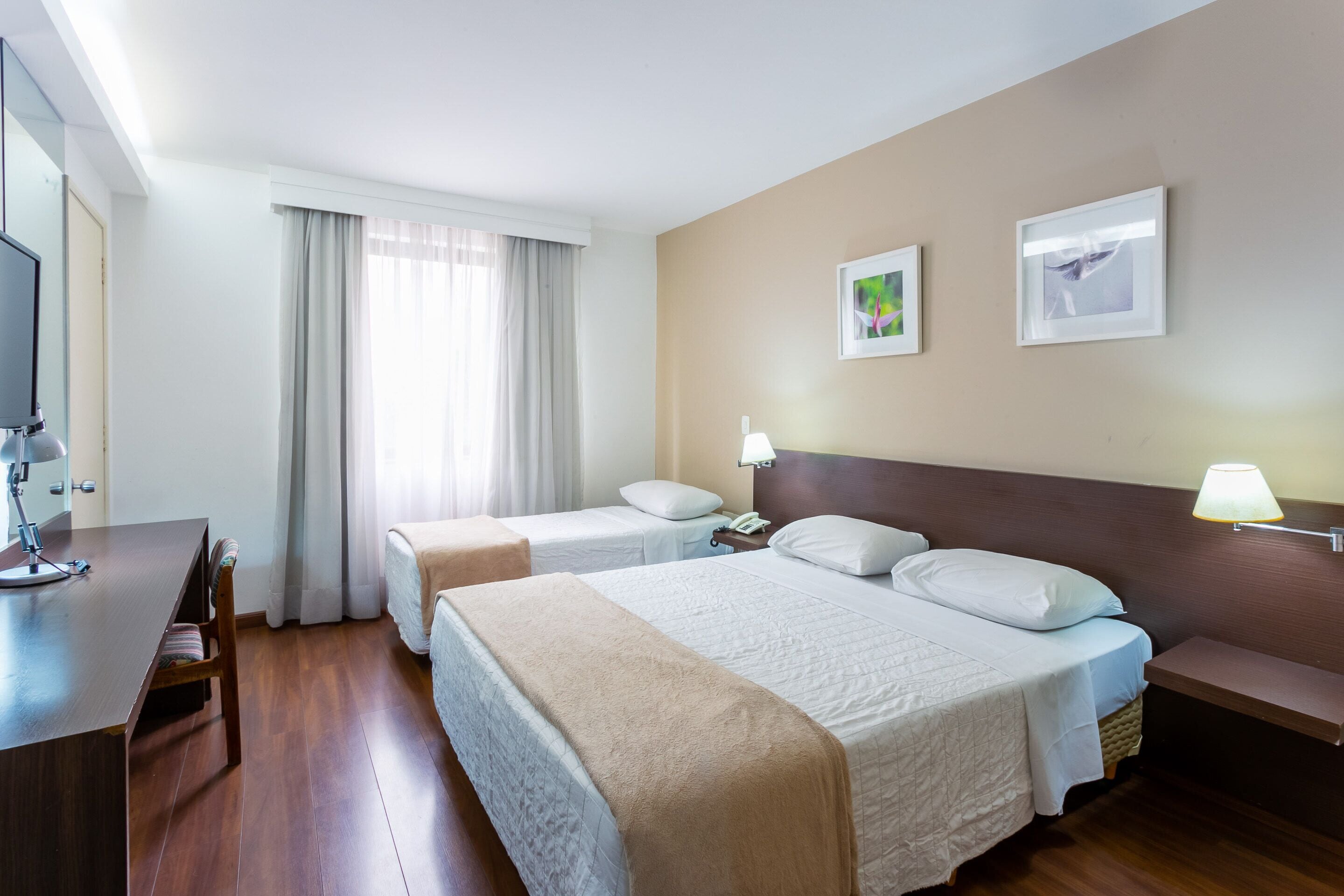 A hotel room with two beds, a desk, a TV, and a window with curtains.