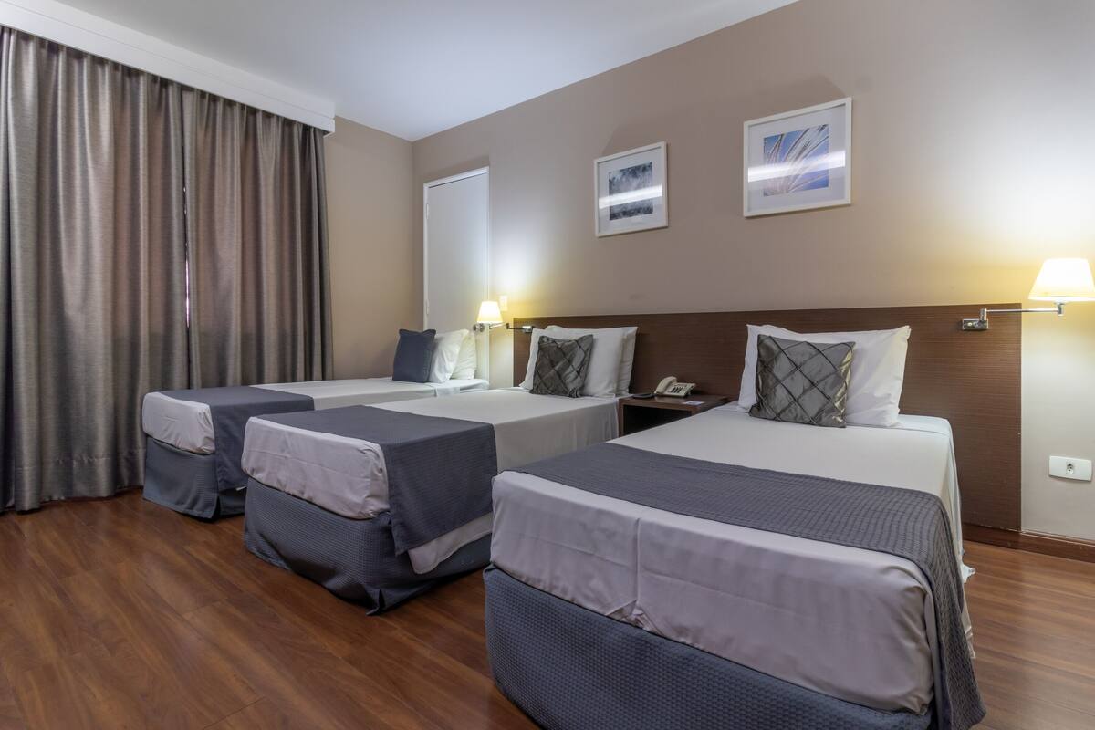 Quarto de hotel com três camas, piso de madeira, uma janela grande com cortinas e duas luminárias fixadas na parede.