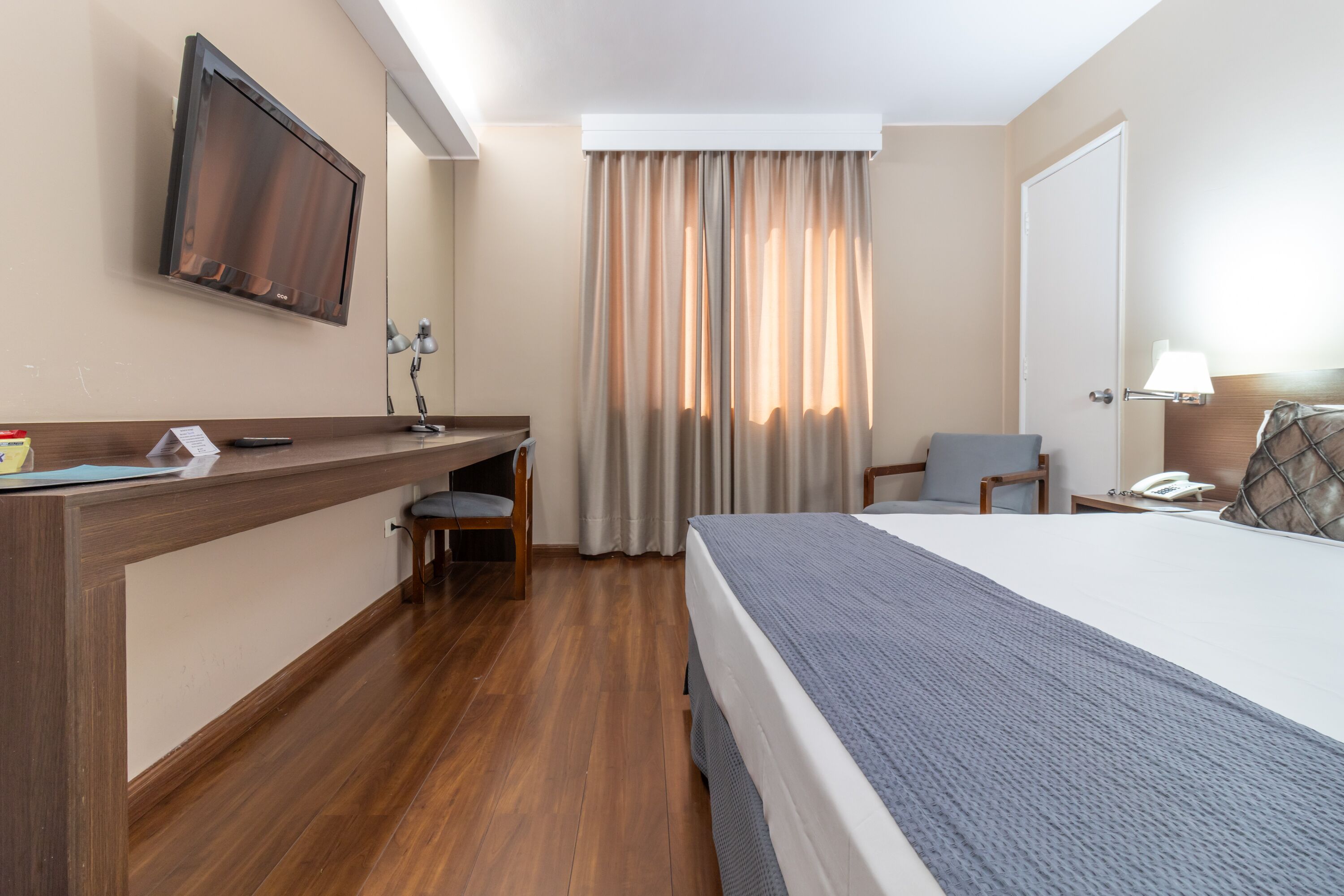 A hotel room with a bed, desk, chair, TV, curtains, and a lamp.