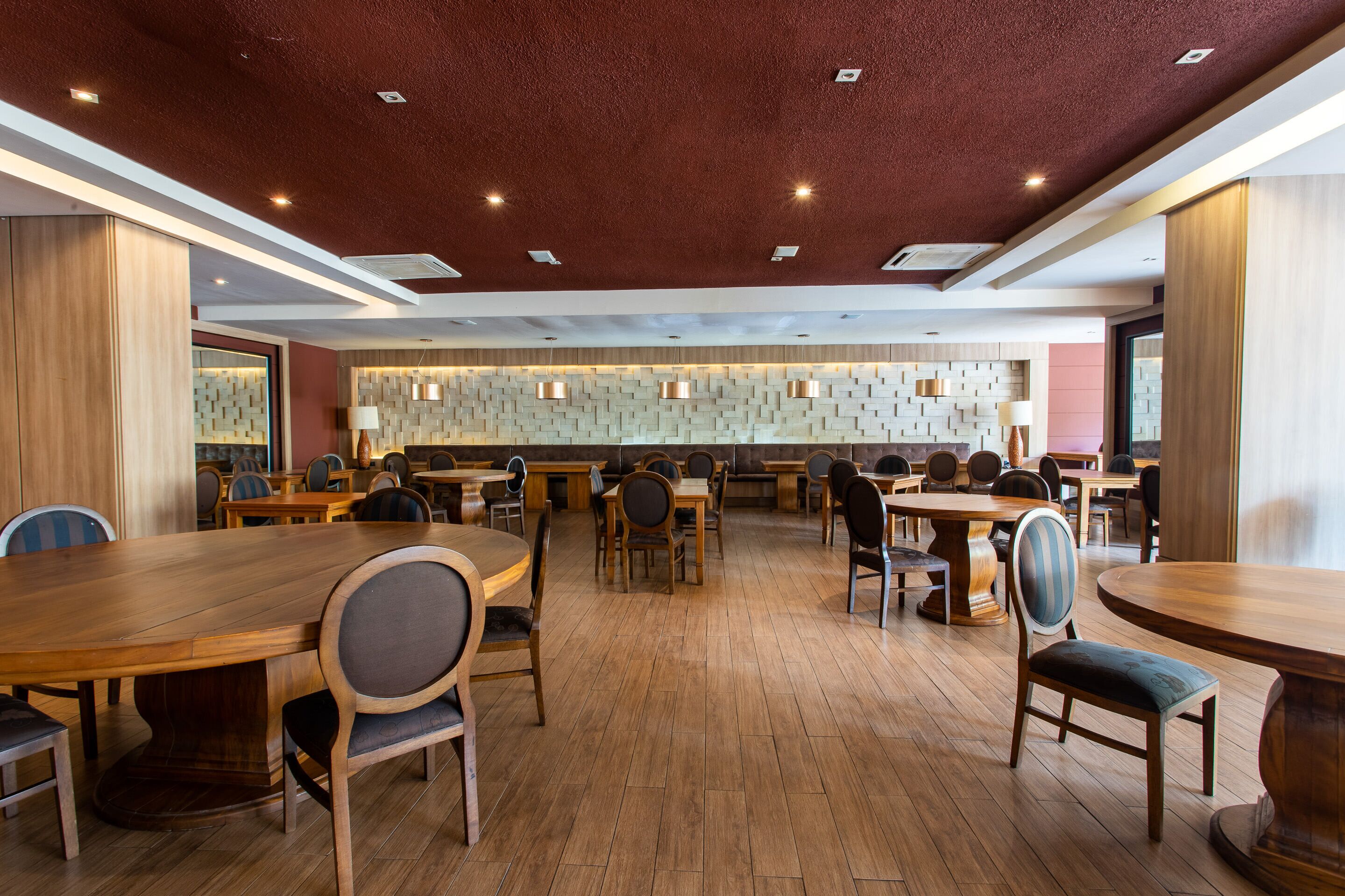 A spacious restaurant with wooden tables and chairs, a patterned wall, and recessed ceiling lighting.