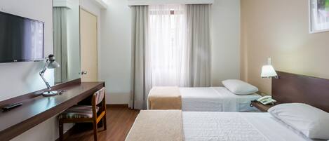 A hotel room with a bed, bedside table, lamp, chair, desk, TV, and a window with curtains.