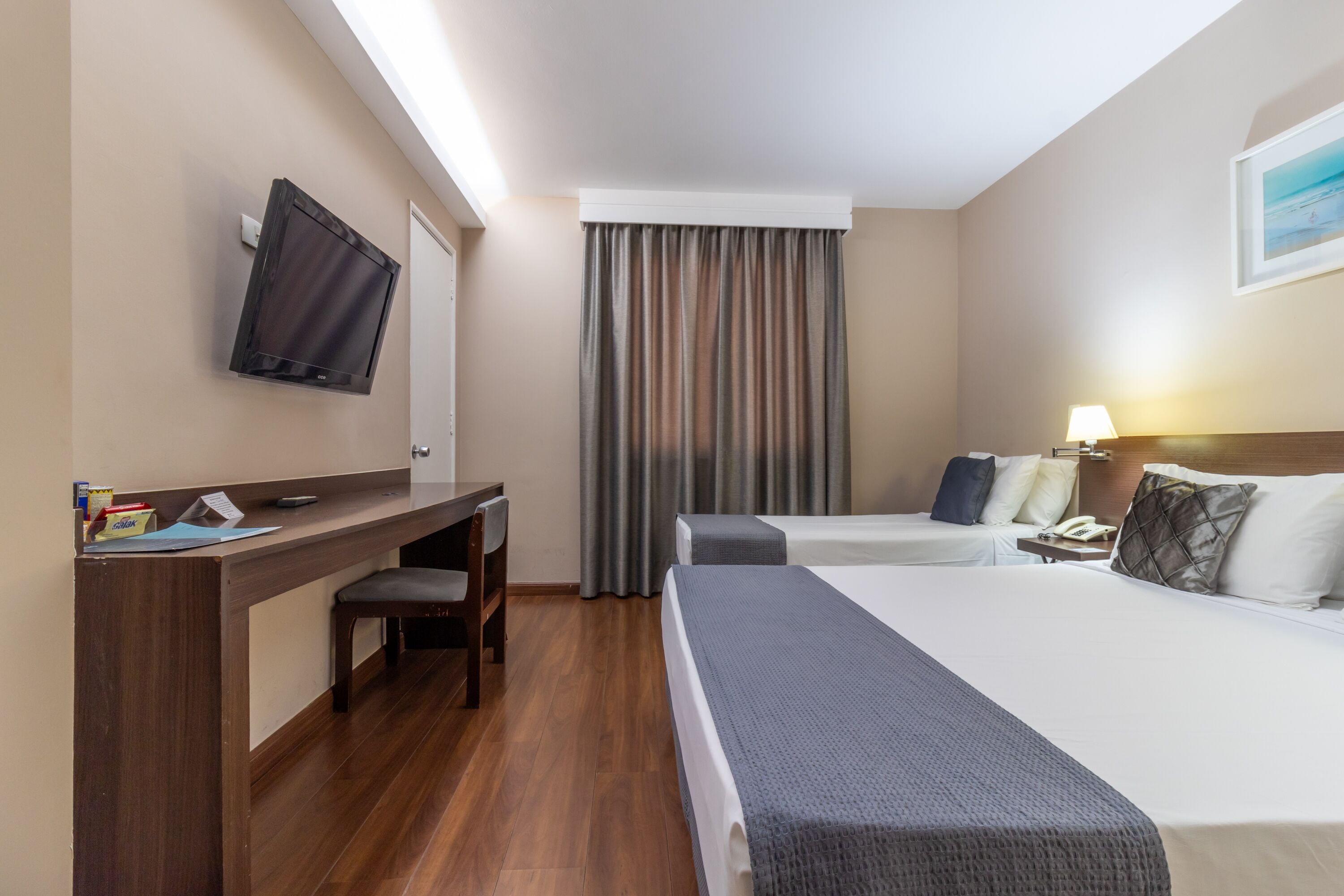 A hotel room with a desk, two beds, a TV, a window with curtains, and a lamp.
