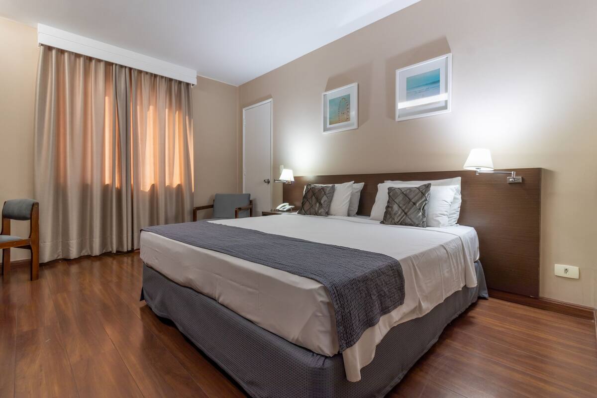 Quarto de hotel com uma cama grande, piso de madeira, uma escrivaninha e uma cadeira.