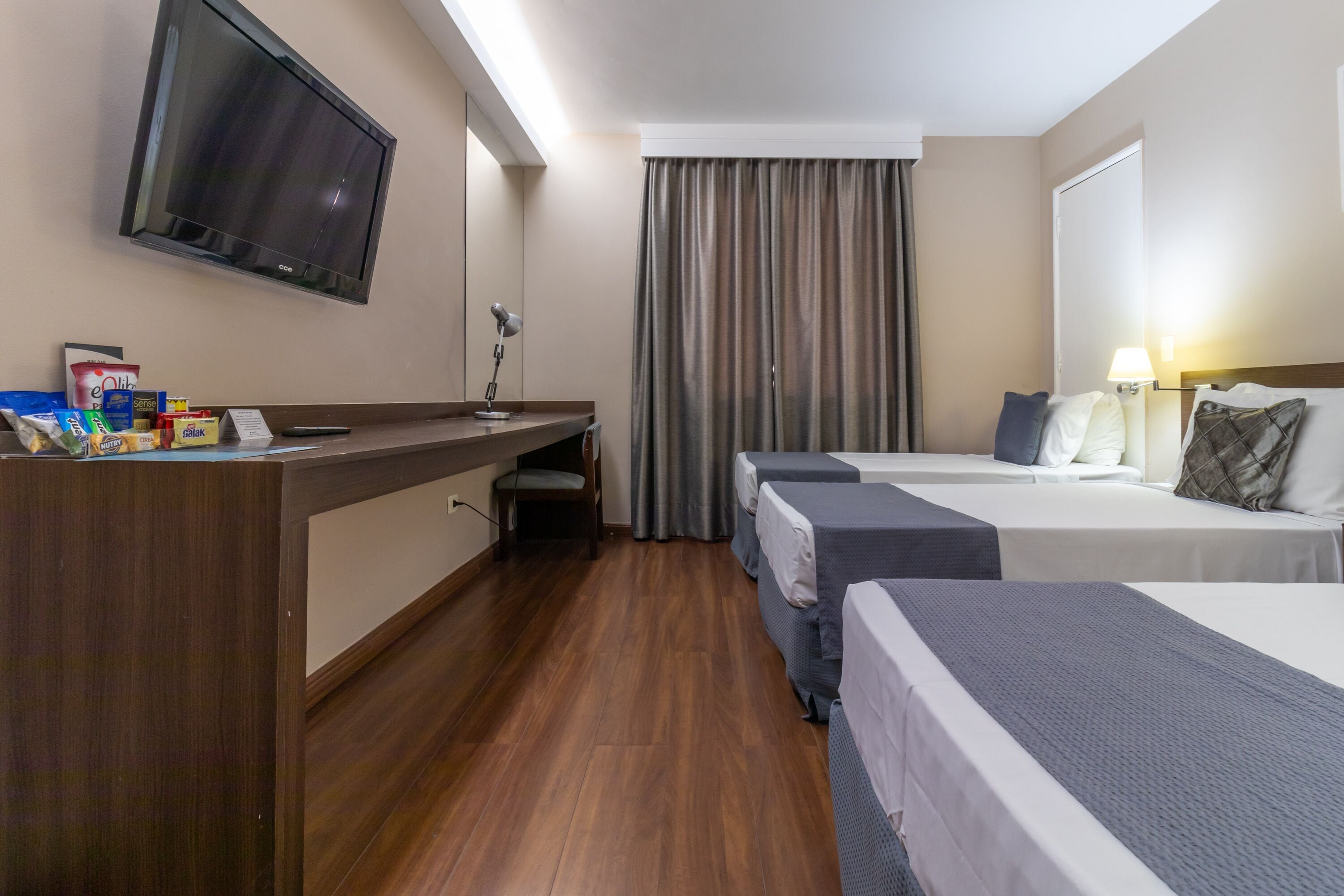 A hotel room with a TV, desk, two beds, a chair, and a curtain.