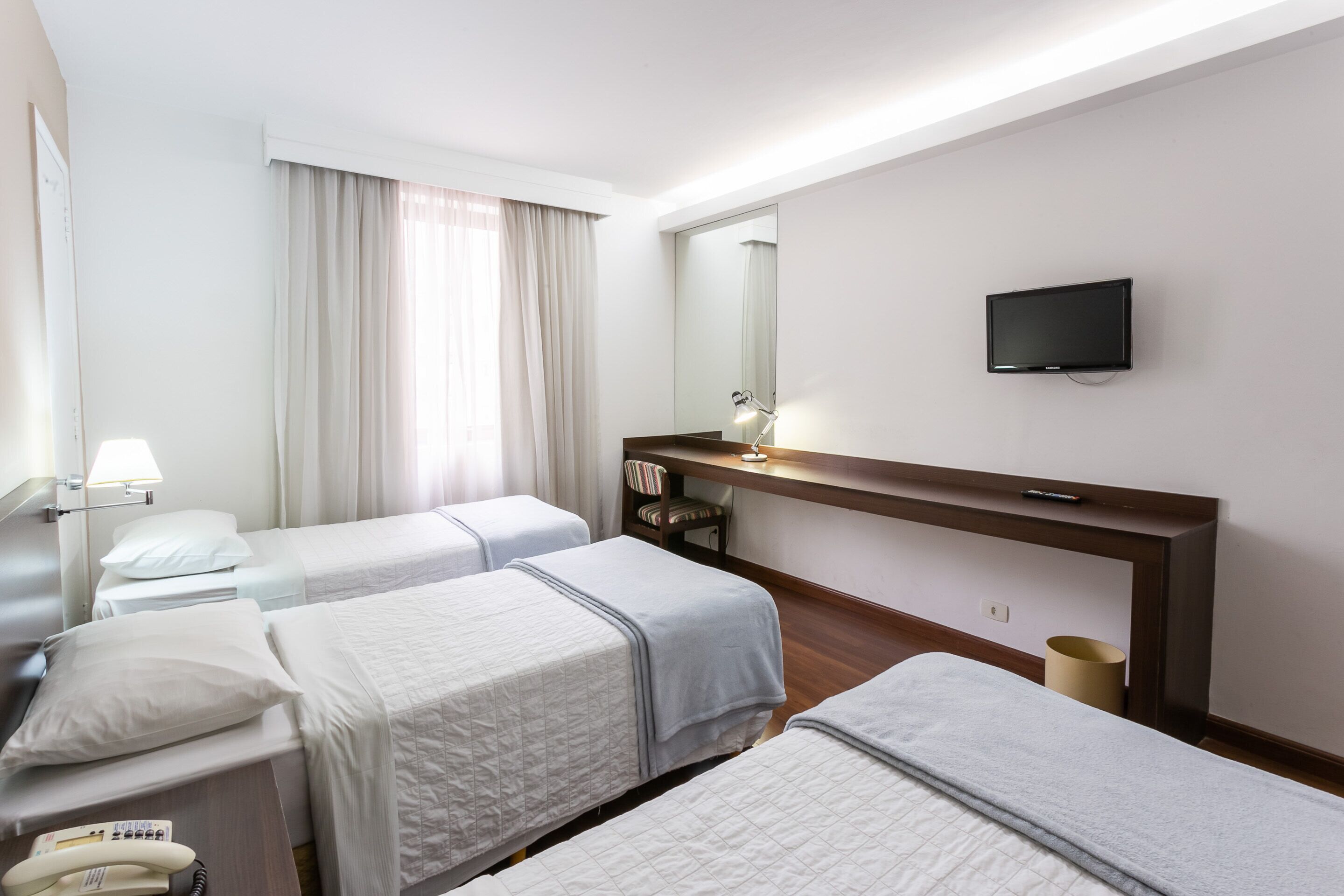 A hotel room with two single beds, a desk with a lamp, a TV mounted on the wall, and a large mirror above the desk.