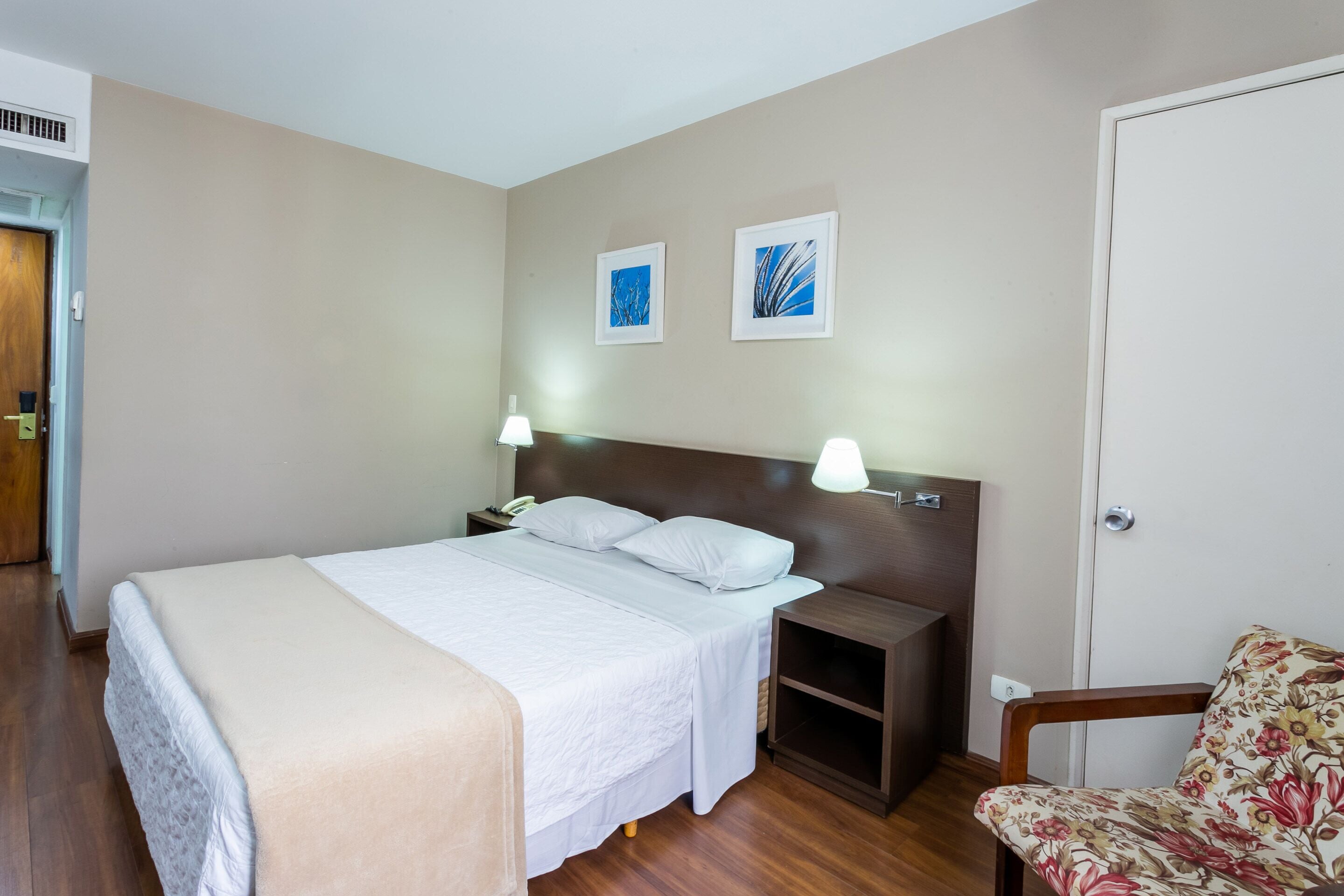 Une chambre d’hôtel avec un grand lit, deux tables de chevet, une petite table, une chaise et deux tableaux encadrés au mur.
