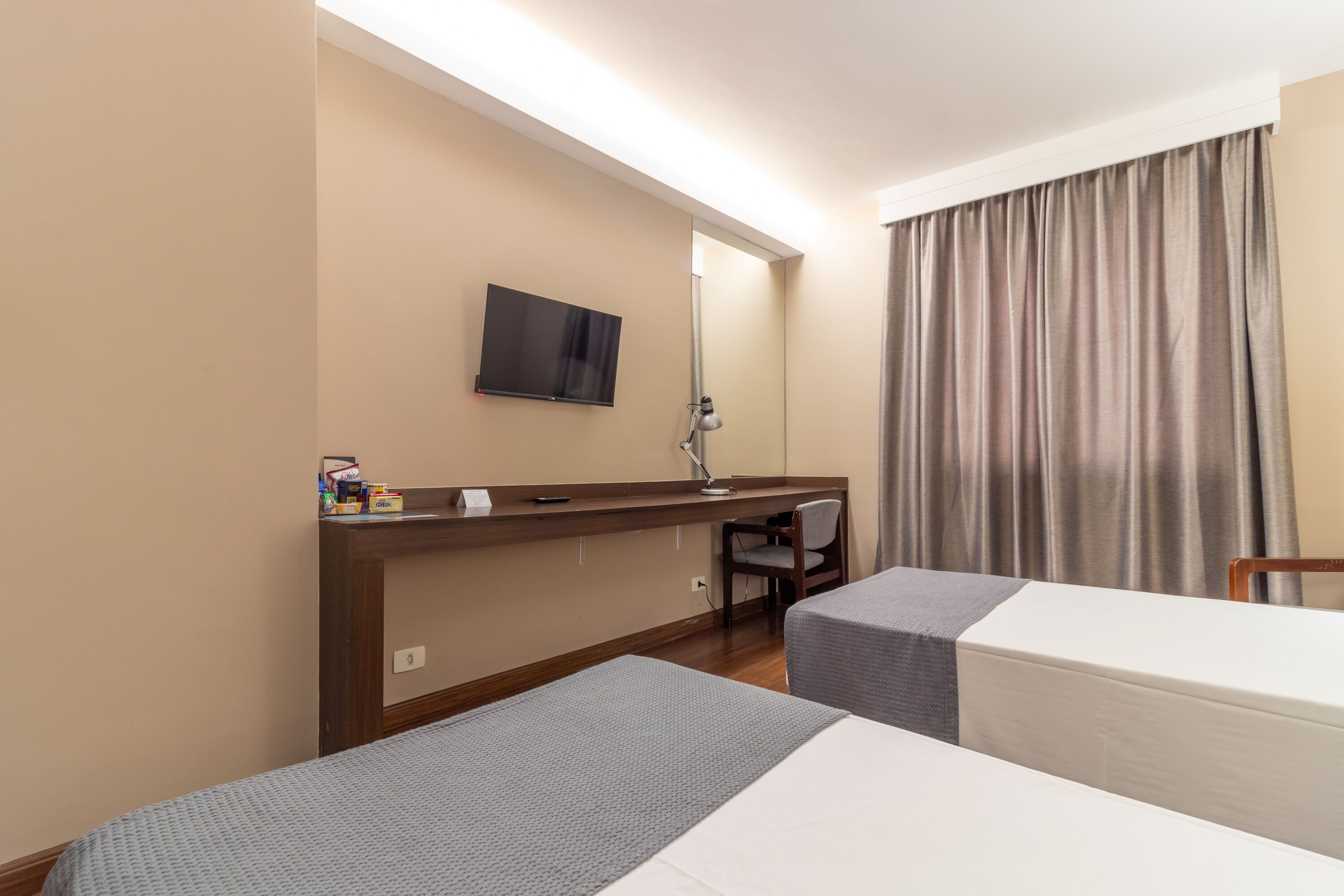 Quarto de hotel com duas camas, uma escrivaninha, uma cadeira, uma TV fixada na parede e uma janela com cortinas.