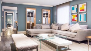 Un salon moderne avec un canapé d’angle, une table basse en verre et un banc. On y trouve deux lampadaires et une œuvre d’art abstraite accrochée au mur.