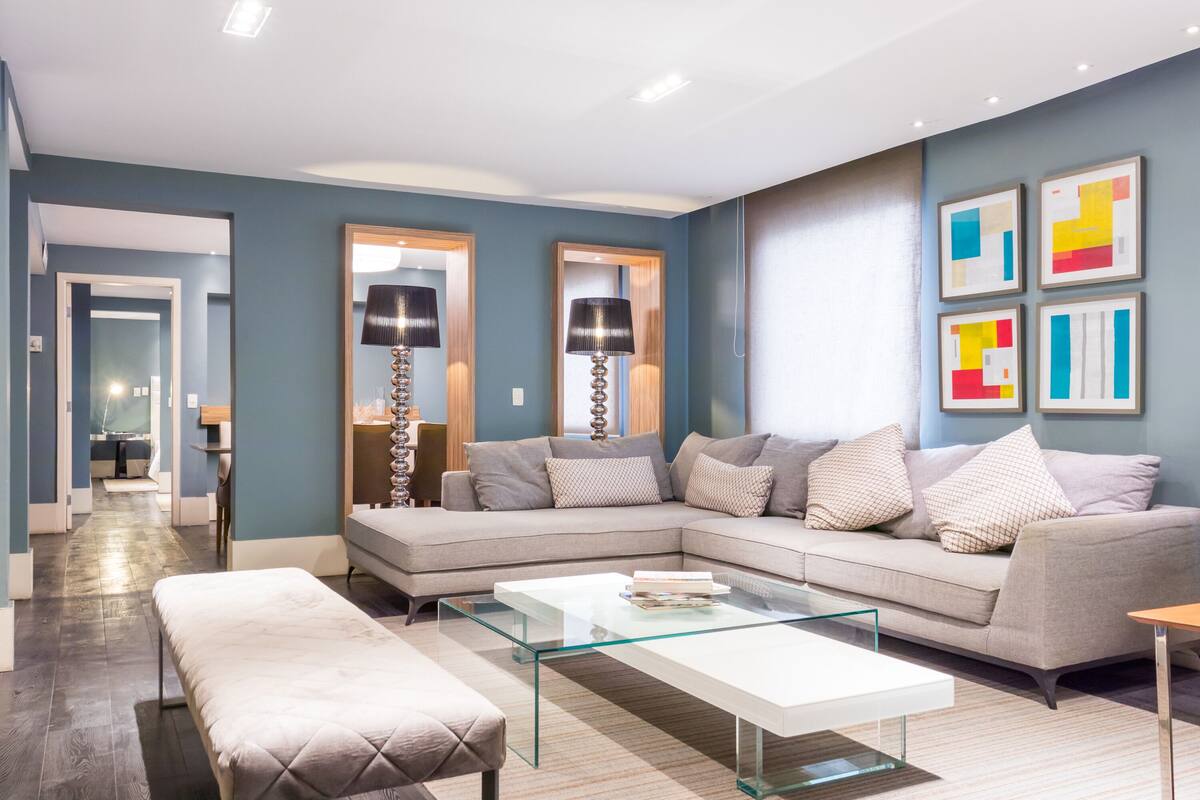 Uma sala de estar moderna com um sofá modular, uma mesa de centro de vidro e um banco. Há duas luminárias de chão e arte abstrata na parede.