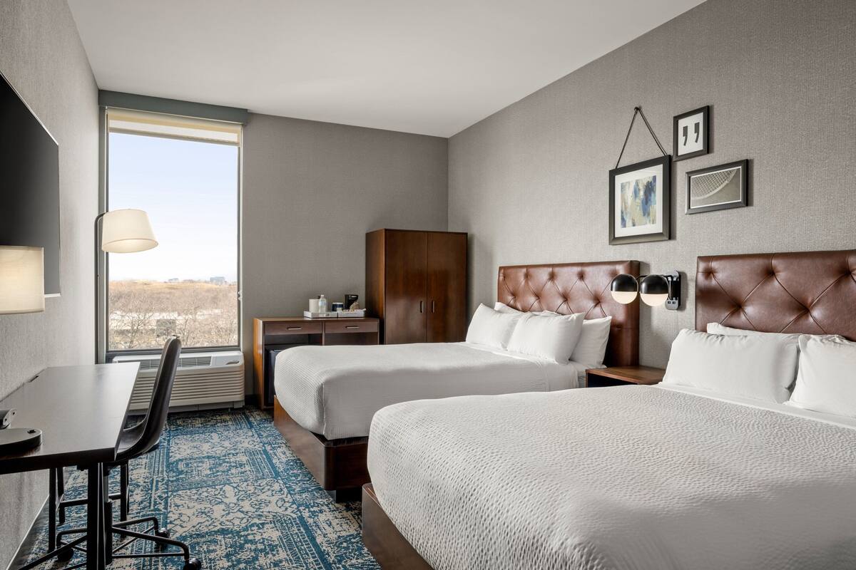 Een hotelkamer met twee bedden, een bureau, een stoel, een kledingkast en een groot raam met uitzicht over de stad.