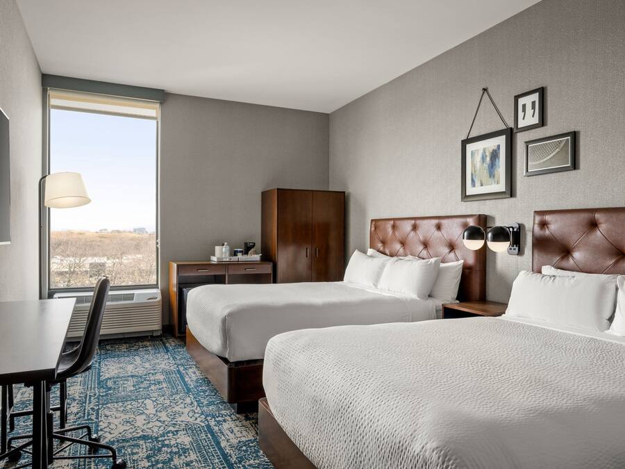 A hotel room with two beds, a desk, a chair, a wardrobe, and a large window offering a city view.