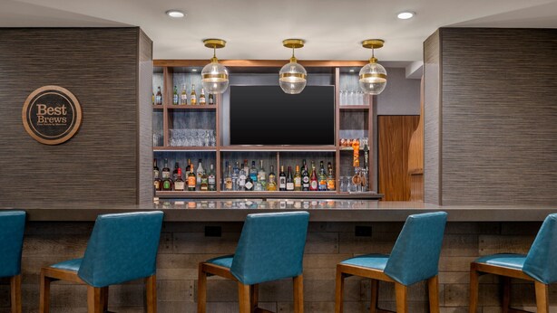 A bar area with a wooden bar counter, blue bar stools, and a variety of liquor bottles. - Four Points by Sheraton Chicago Westchester/Oak Brook (Westchester)